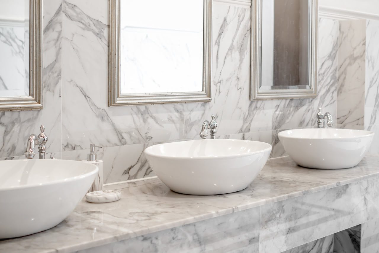 Modern bathroom with three white vessel sinks on a marble countertop, featuring silver faucets and framed mirrors above each sink.