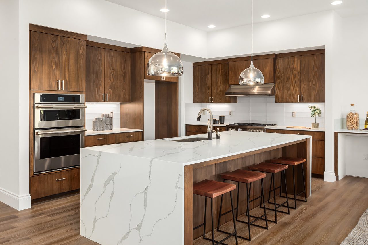 Modern kitchen with wooden cabinets, a large marble island with sink, and four bar stools. Pendant lights hang above the island, and stainless steel appliances are in the background.