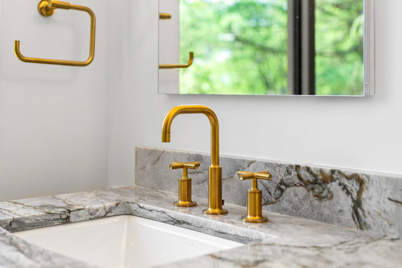 A bathroom sink with a marble countertop, a gold faucet set, and a mirror above, reflecting green foliage outside.