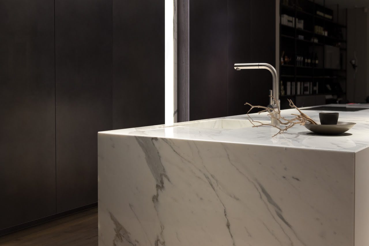 Minimalist kitchen with a marble island countertop, a sleek faucet, and a dark back wall. A small branch and cup rest on the counter.