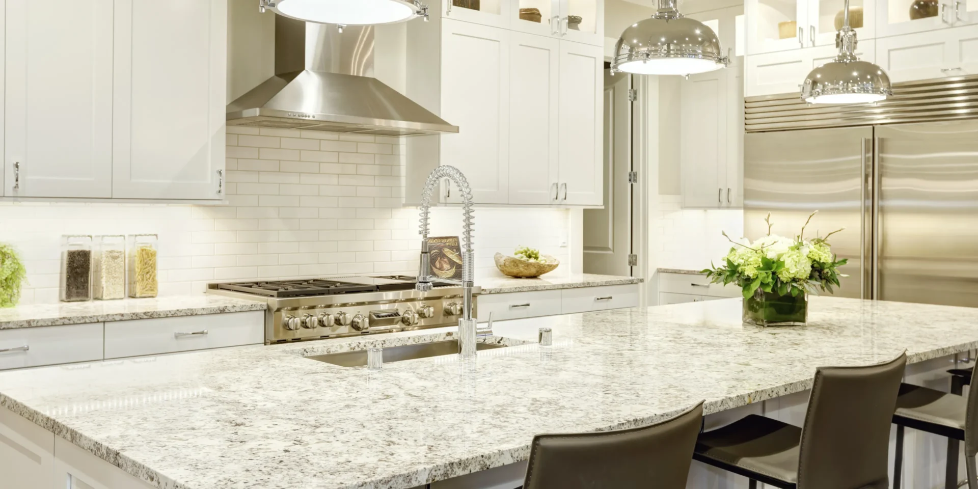 Modern kitchen with white cabinets, stainless steel appliances, and granite countertop. Large island with seating, overhead pendant lights, and decorative plants are present.