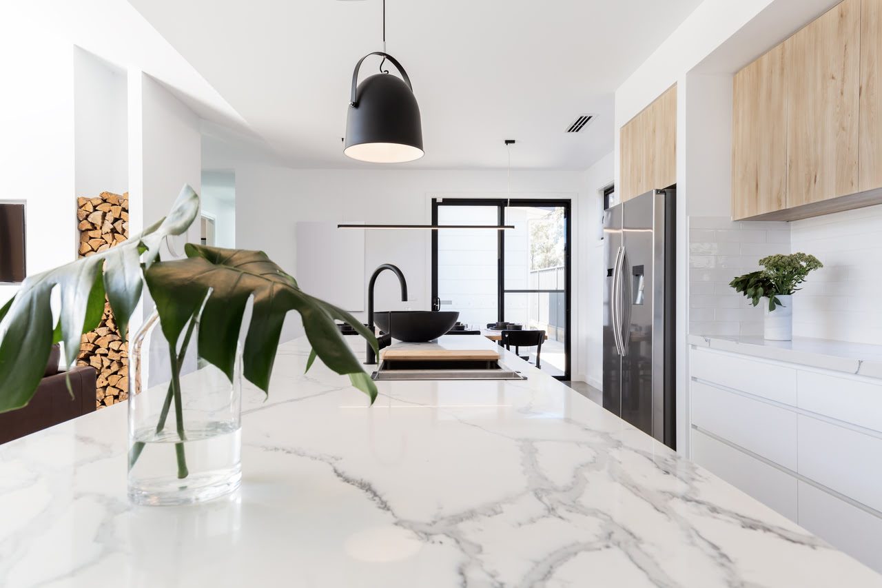 Modern kitchen with a marble countertop, pendant lights, and wooden cabinets. A glass vase with a large leaf is on the counter. The room is bright and spacious.